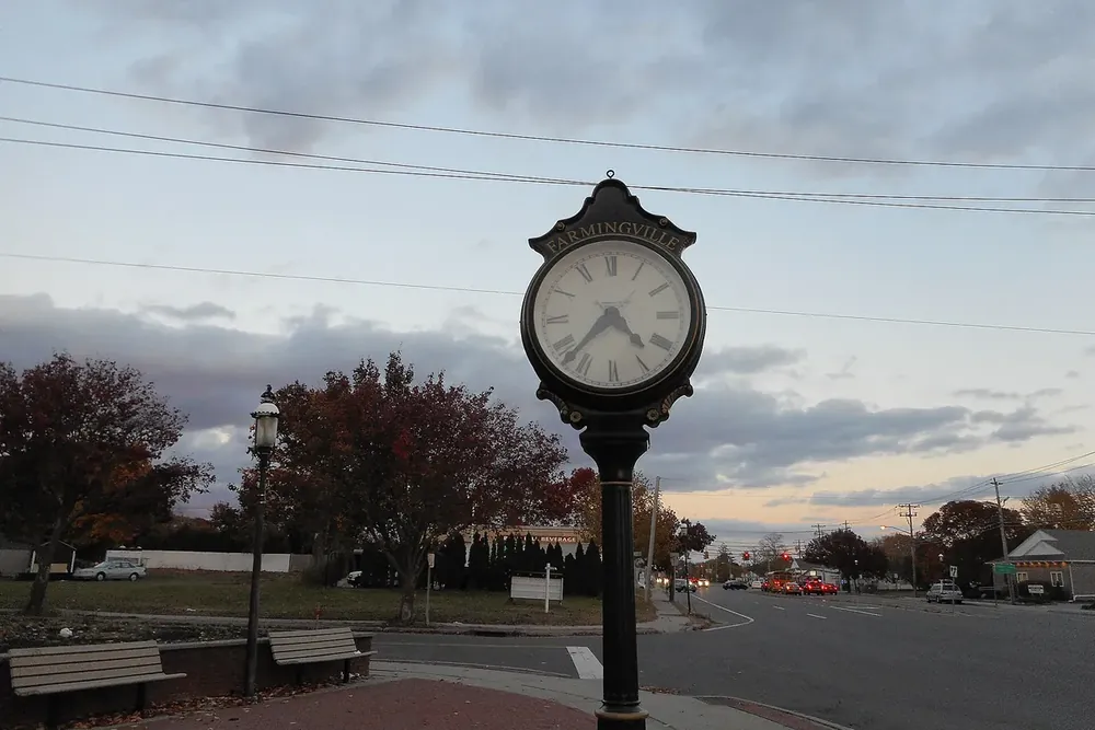 Tall sidewalk street clock beside benches on a corner at dusk in Farmingville, New York