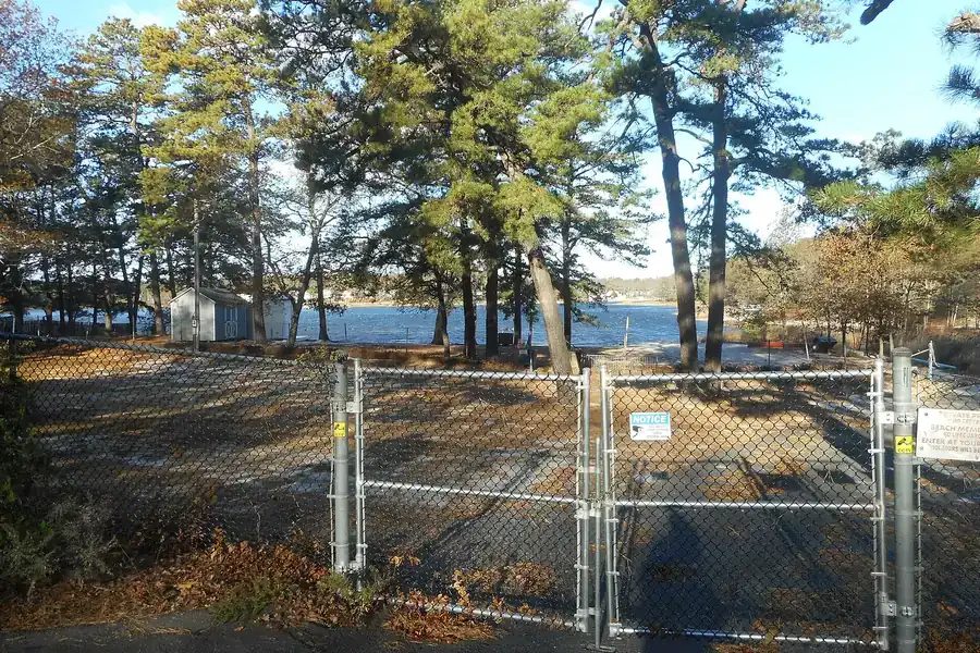 Chain-link gate beside a pine-lined lake shore at Lake Panamoka Beach in Ridge, New York